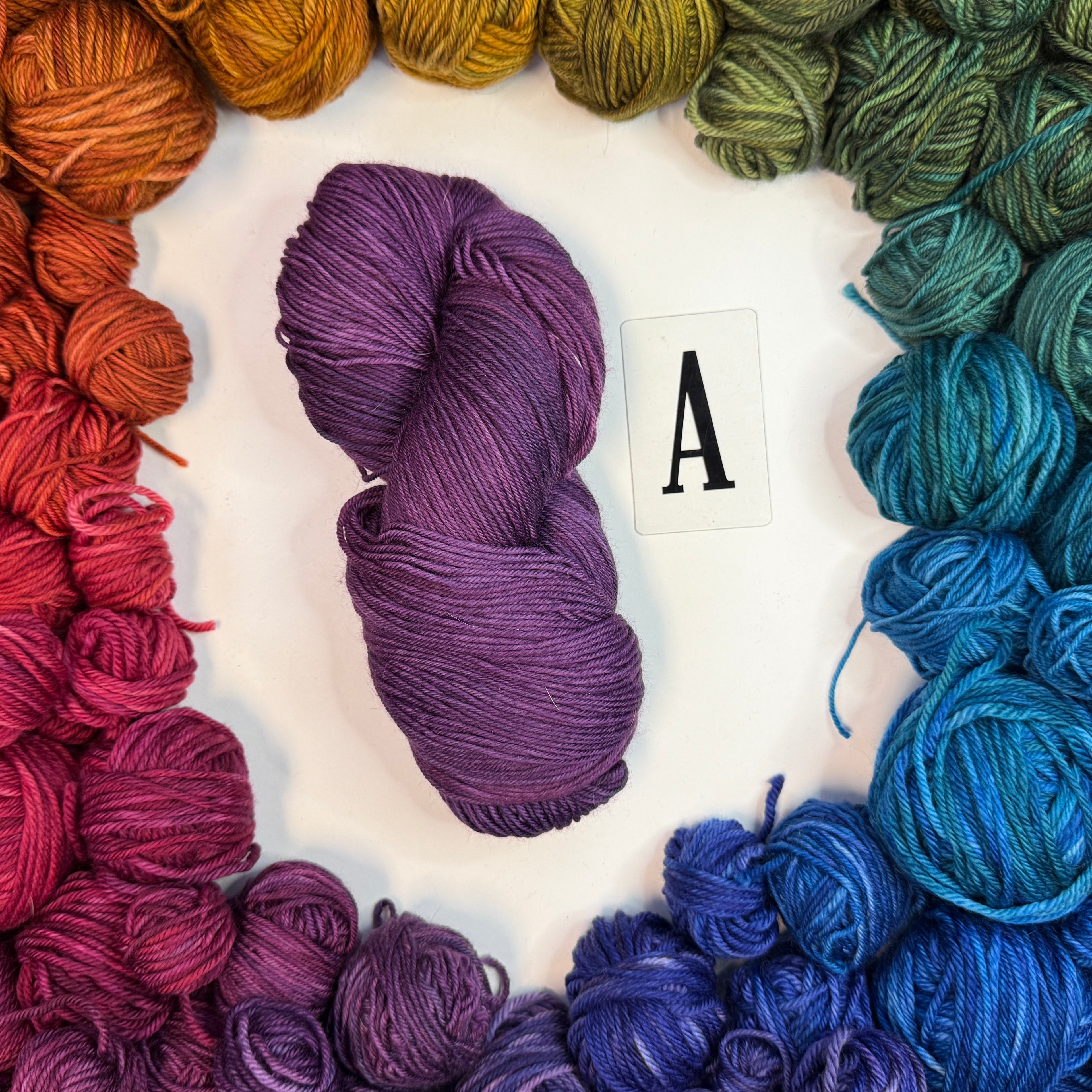 A purple skein of yarn labeled "A" framed by a circle of a variety of balls of yarn in rainbow order.