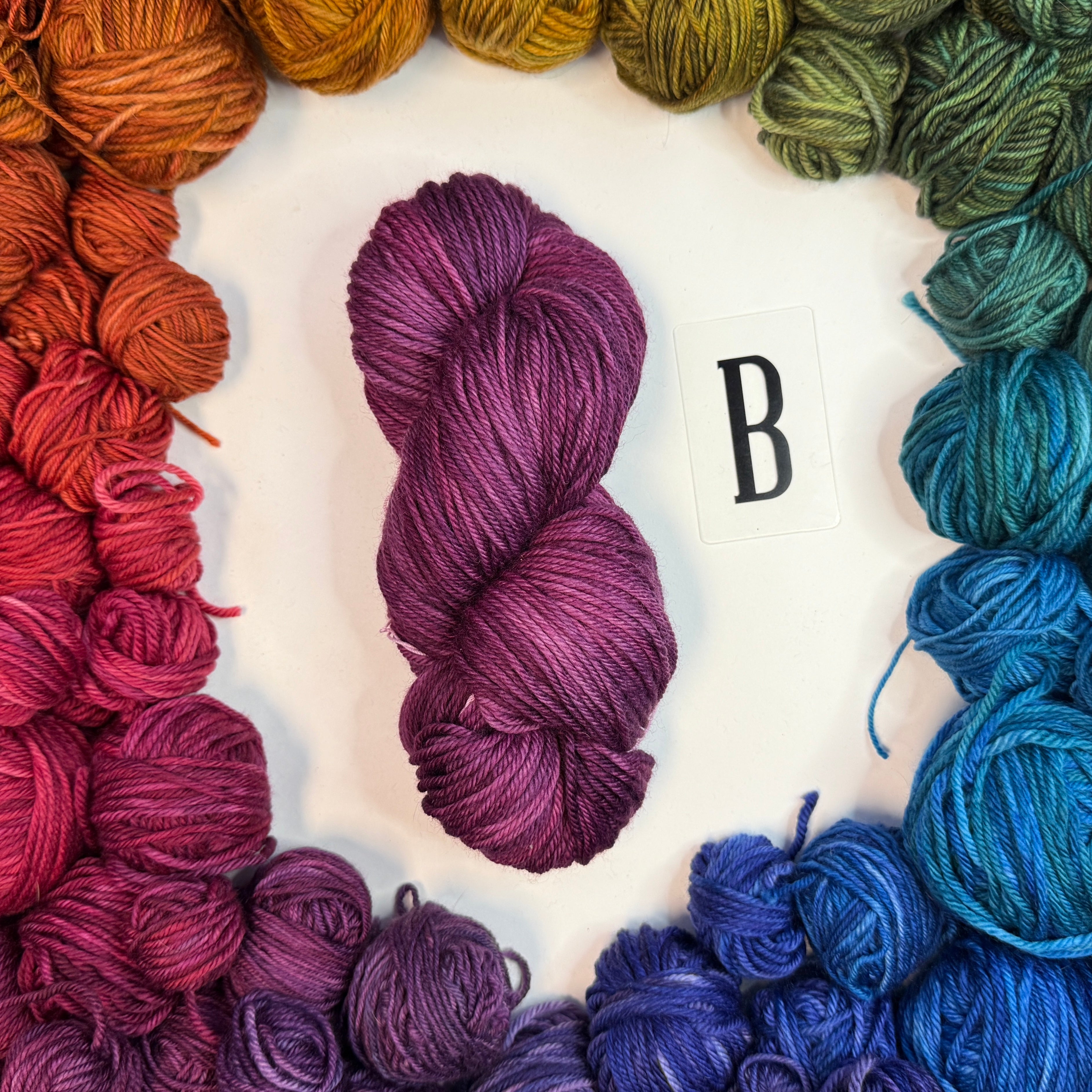 A magenta skein of yarn labeled "B" framed by a circle of a variety of balls of yarn in rainbow order.