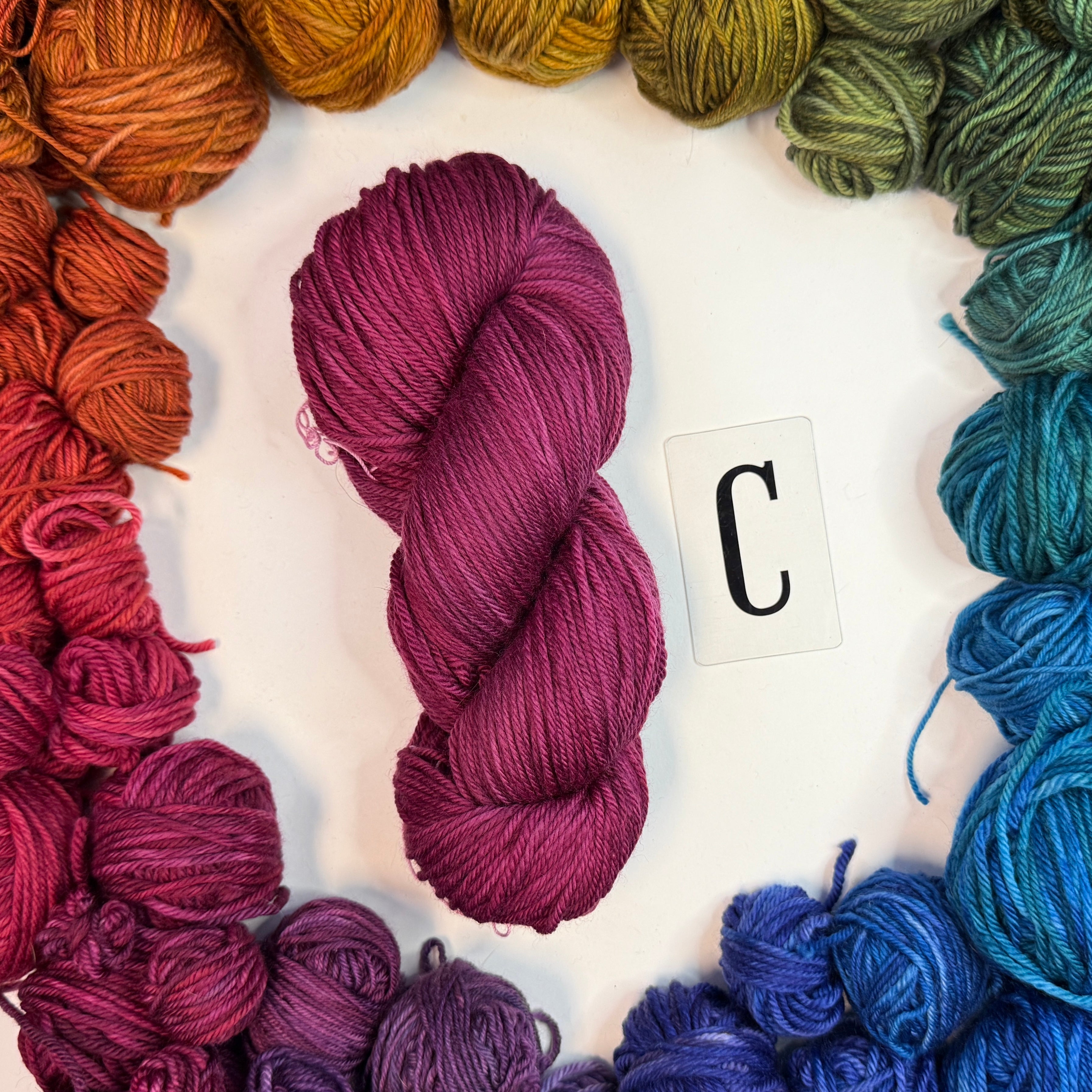A dark pink skein of yarn labeled "C" framed by a circle of a variety of balls of yarn in rainbow order.