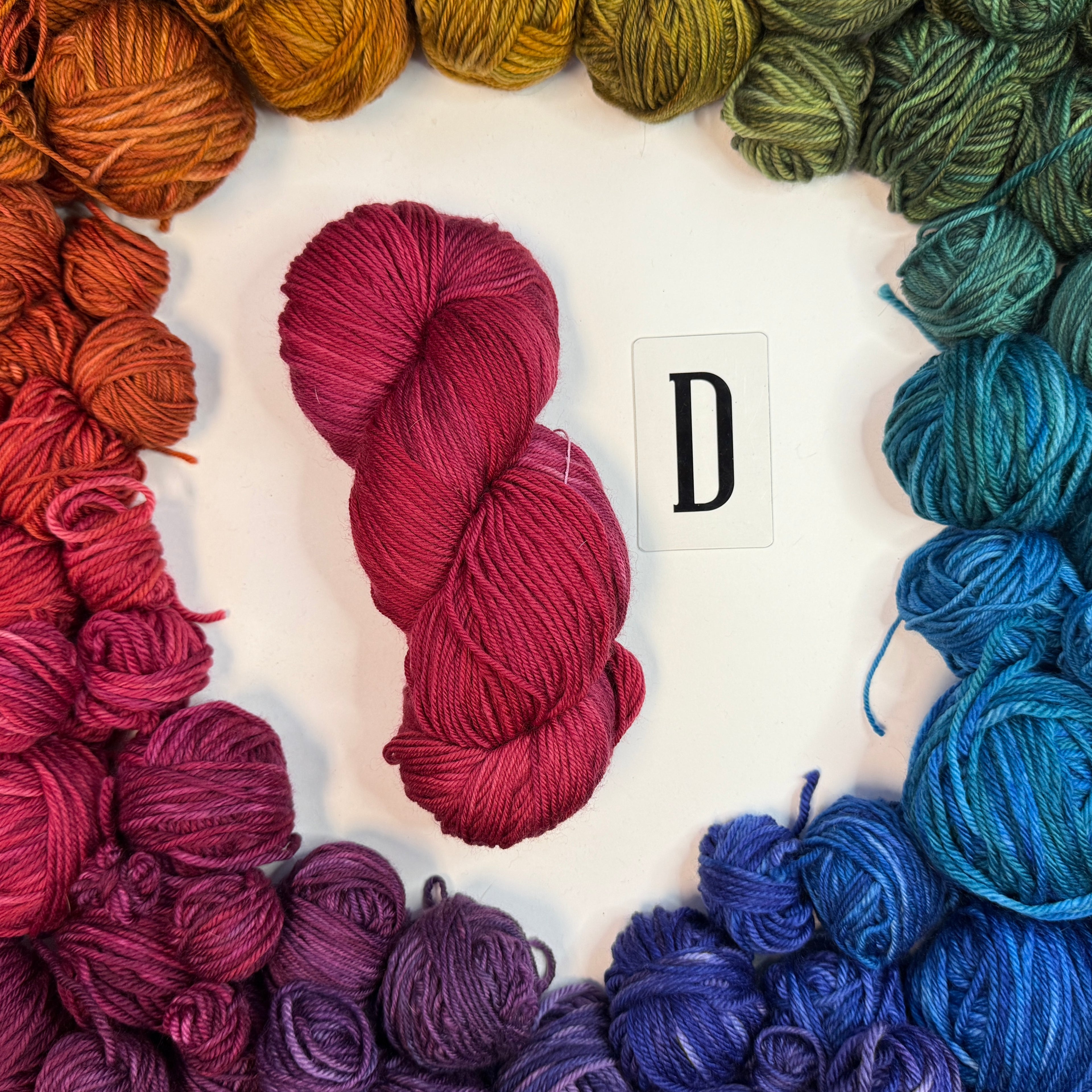 A red skein of yarn labeled "D" framed by a circle of a variety of balls of yarn in rainbow order.