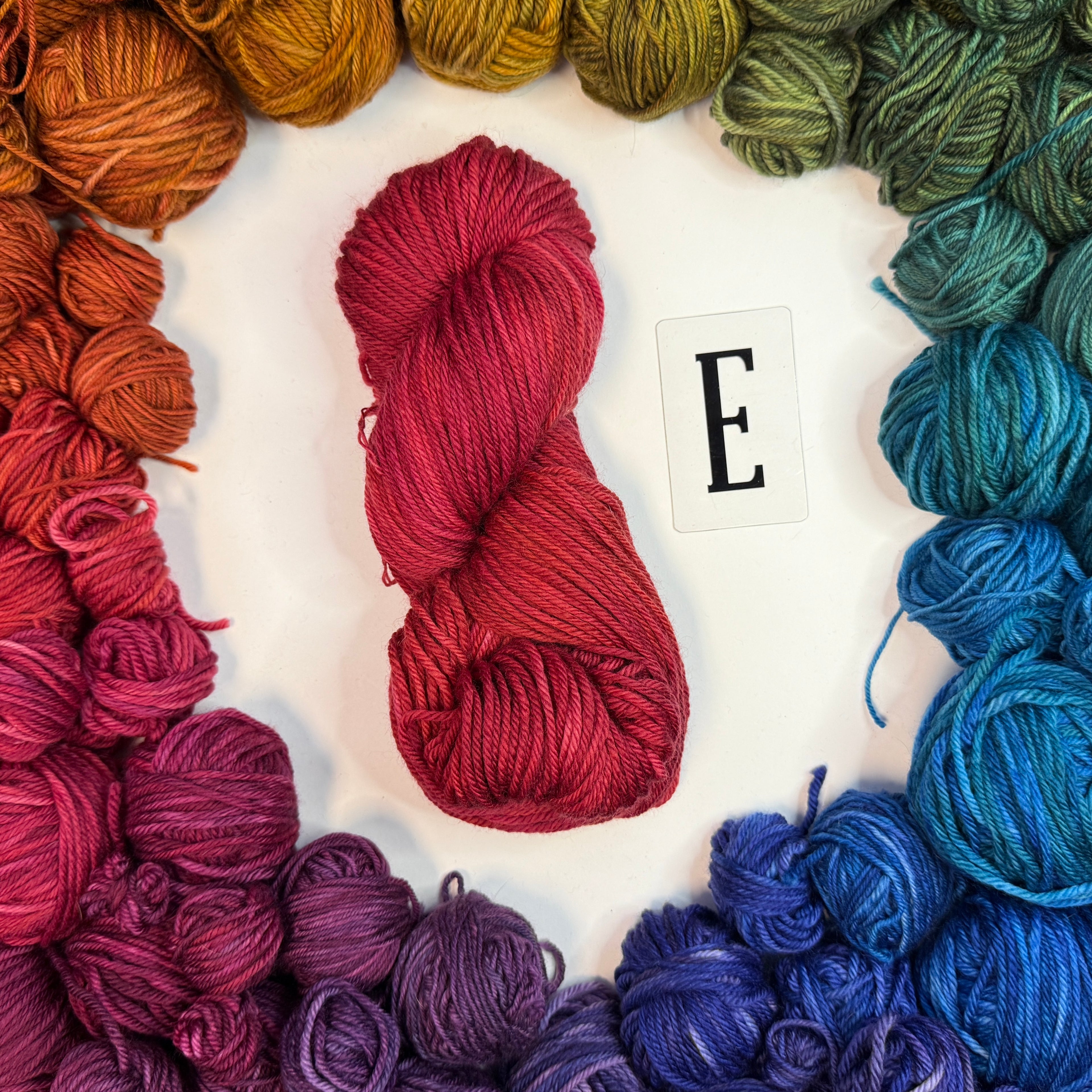 A red skein of yarn labeled "E" framed by a circle of a variety of balls of yarn in rainbow order.