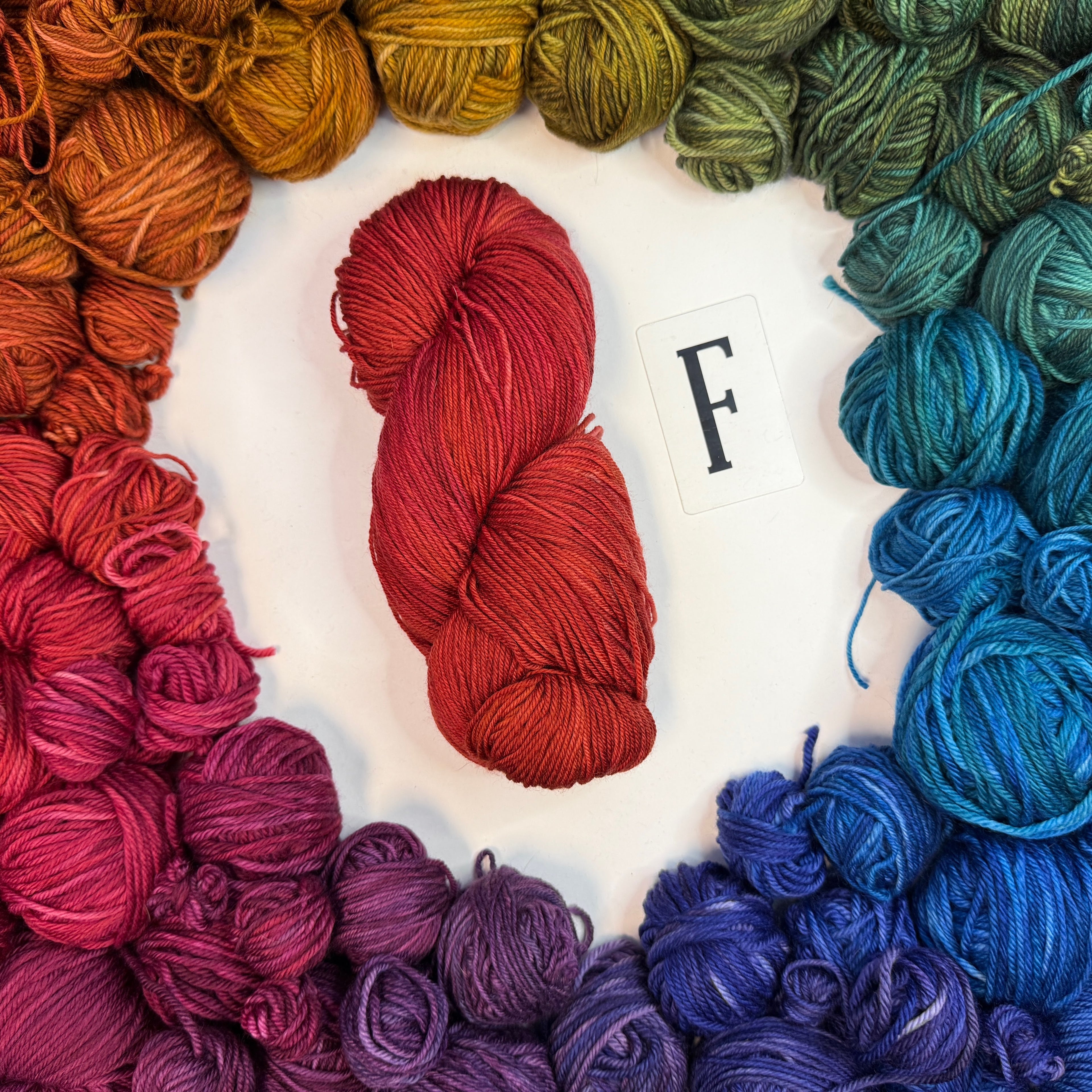 Red-Orange skein of yarn labeled F, surrounded by a circle of yarn balls in  colors in color wheel order.