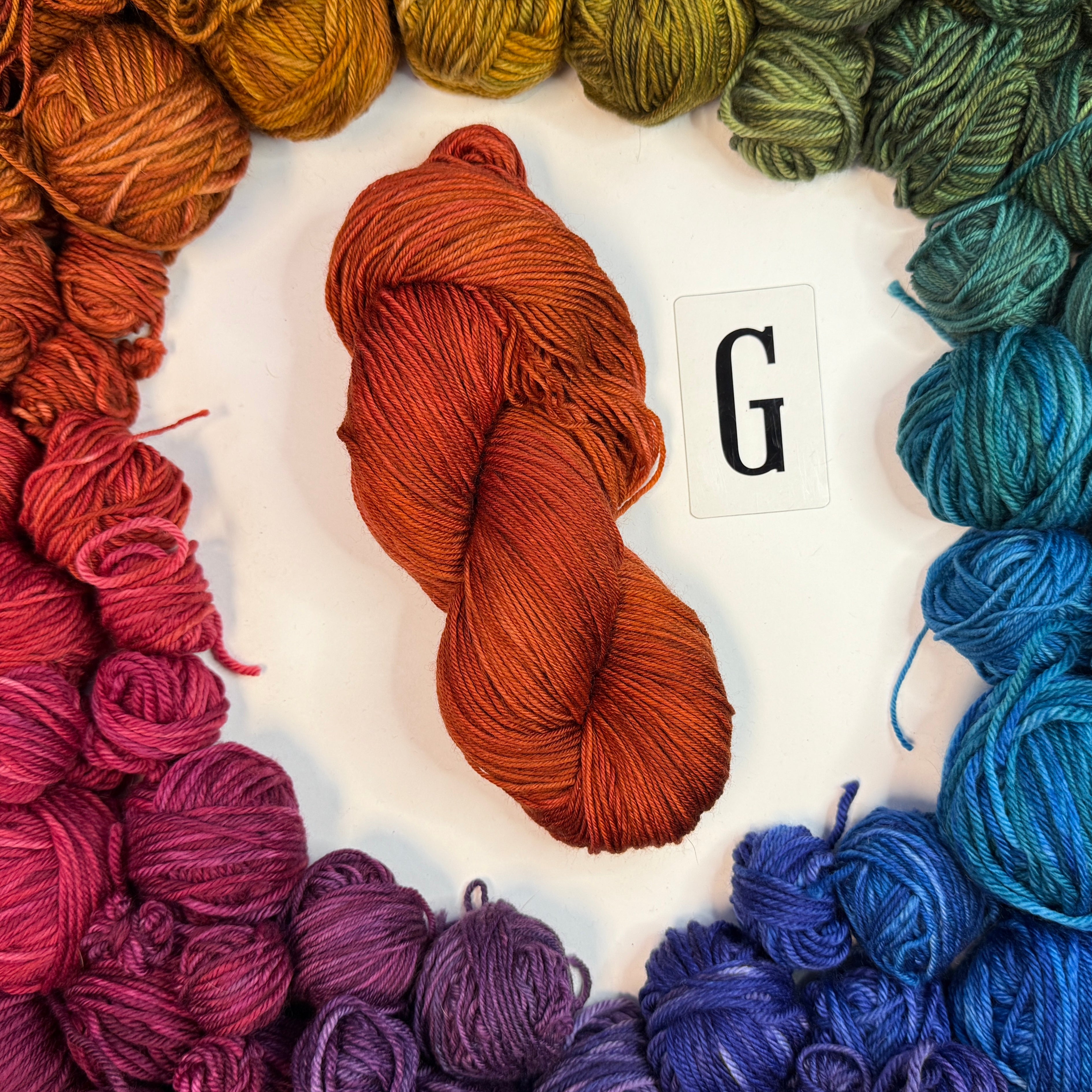 An orange skein of yarn labeled "G" framed by a circle of a variety of balls of yarn in rainbow order.