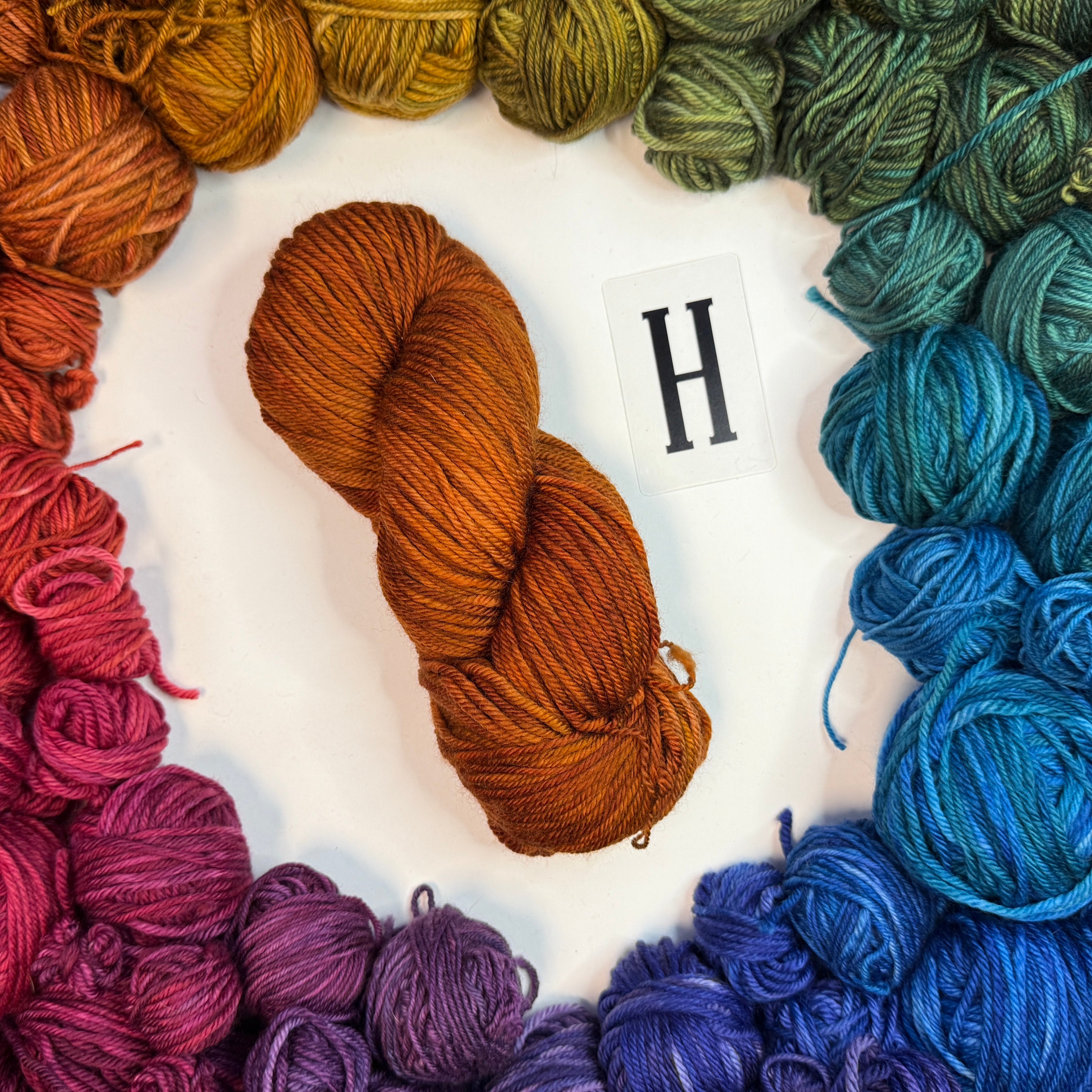 An orange skein of yarn labeled "H" framed by a circle of a variety of balls of yarn in rainbow order.