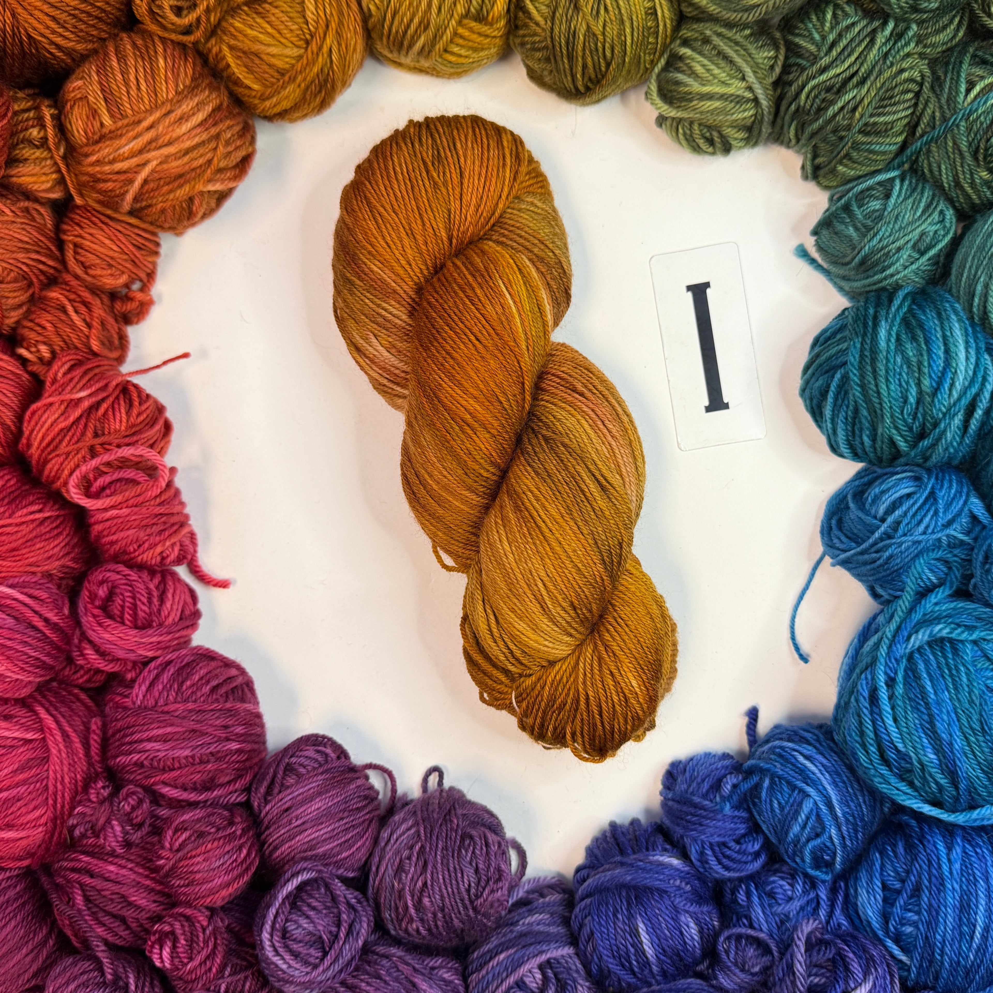A yellow-orange skein of yarn labeled "I" framed by a circle of a variety of balls of yarn in rainbow order.