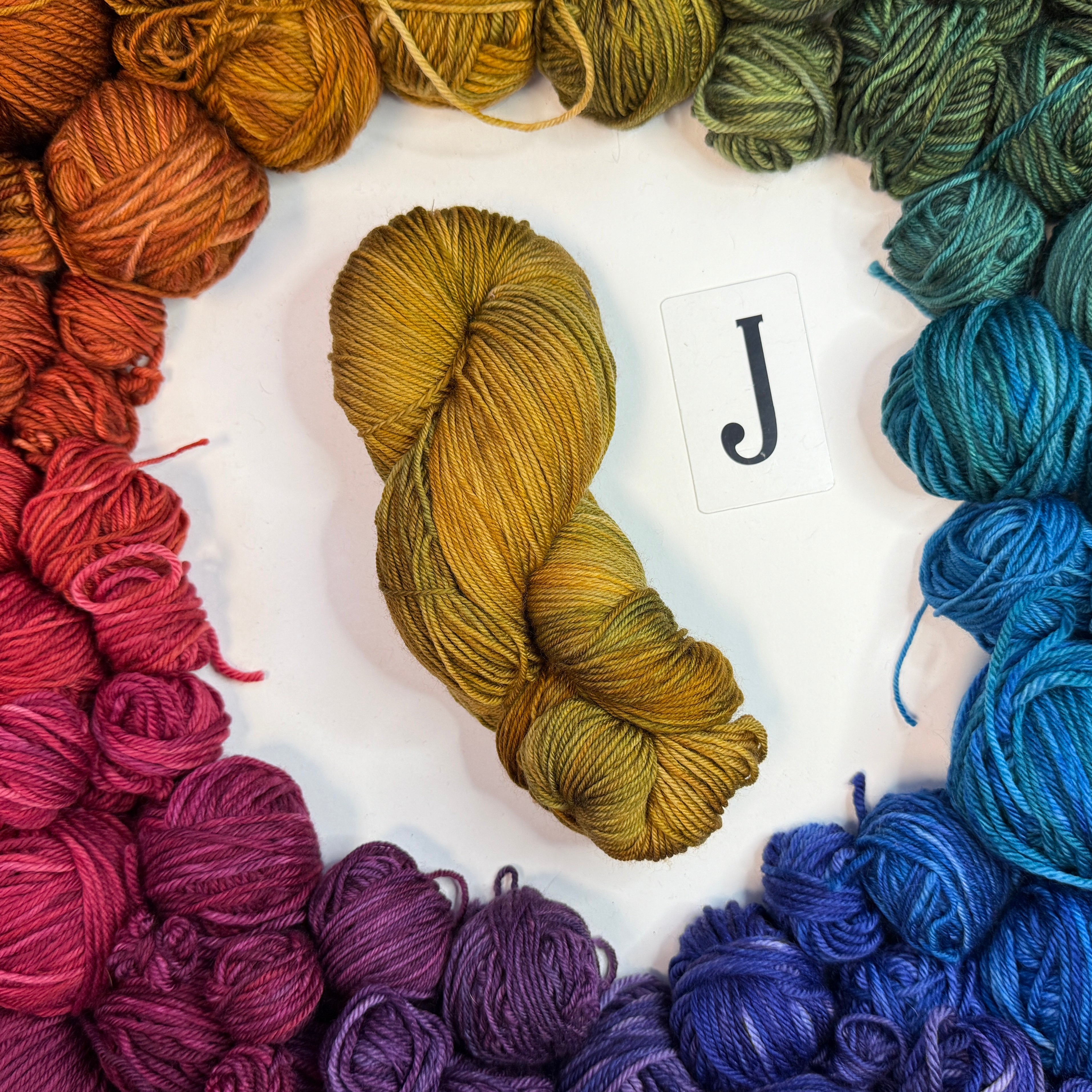 A yellowish skein of yarn labeled "J" framed by a circle of a variety of balls of yarn in rainbow order.