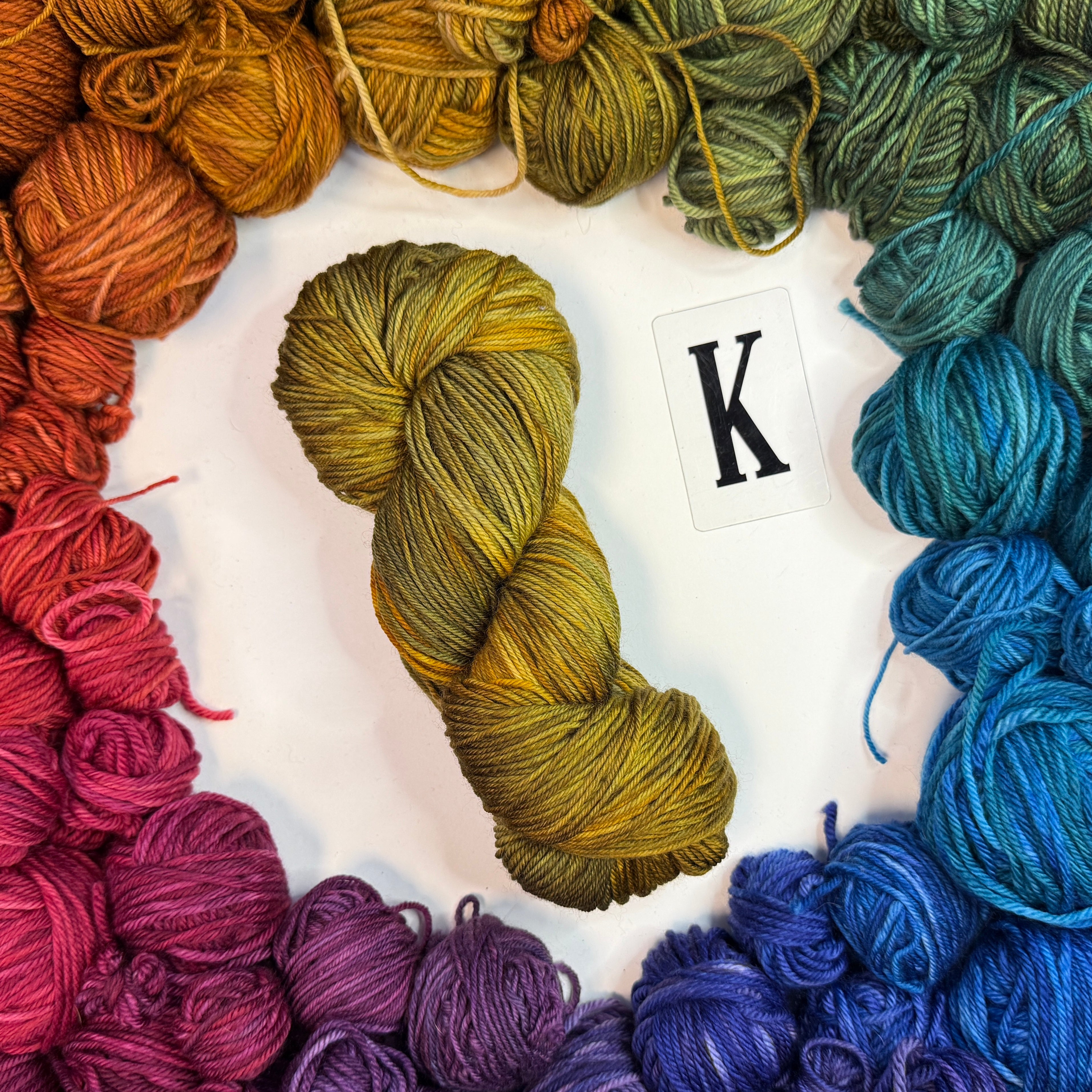 A yellowish skein of yarn labeled "K" framed by a circle of a variety of balls of yarn in rainbow order.