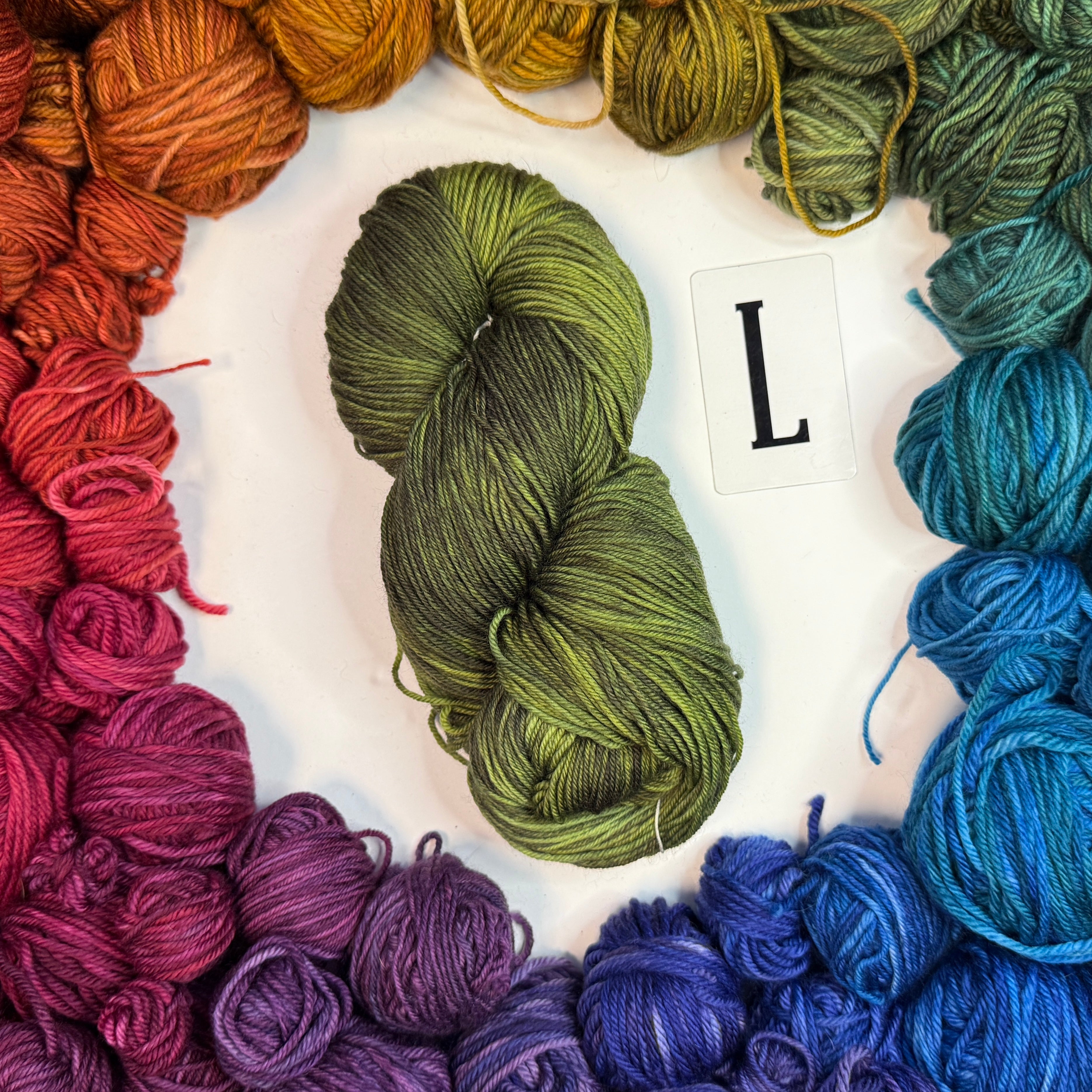 A green skein of yarn labeled "L" framed by a circle of a variety of balls of yarn in rainbow order.