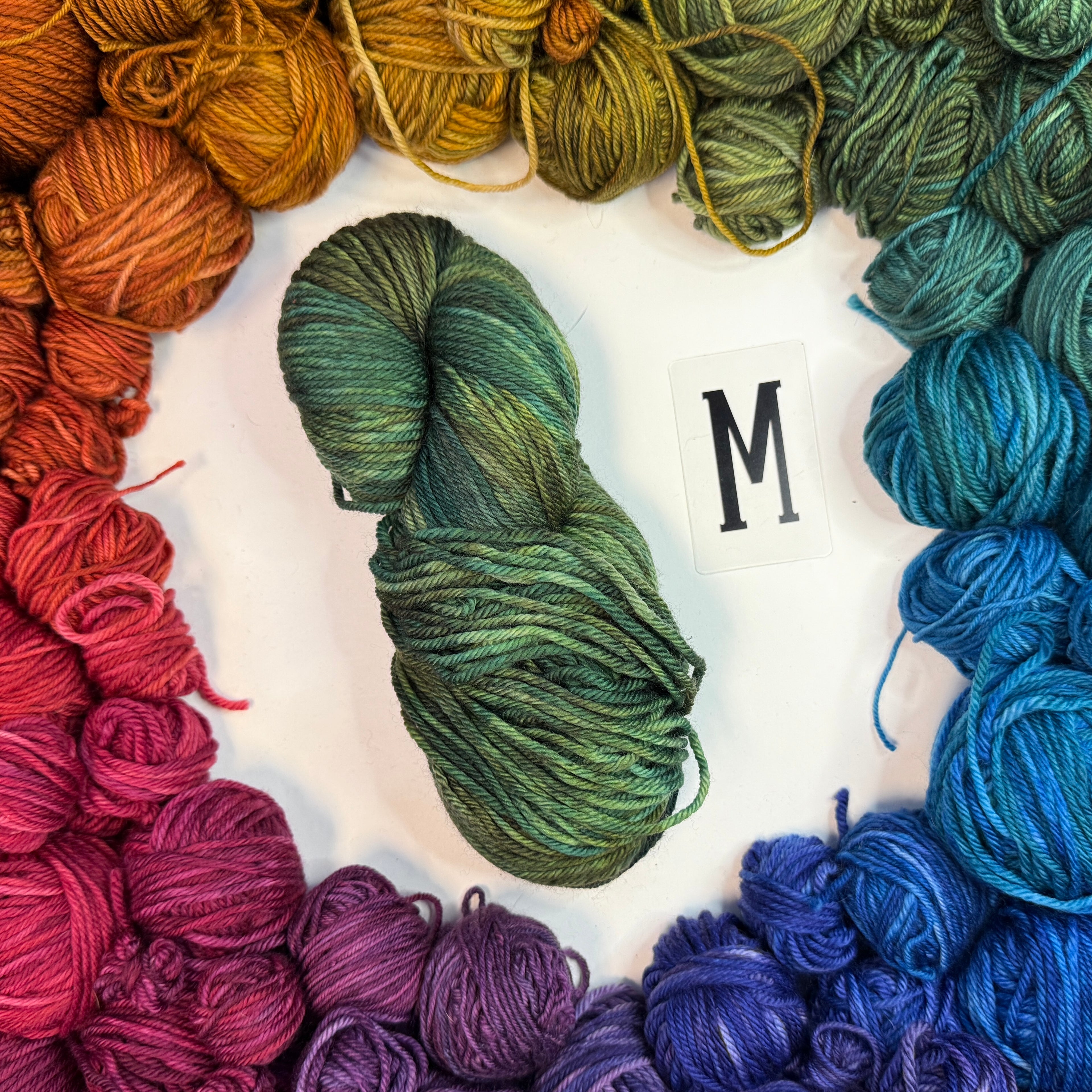 A green skein of yarn labeled "M" framed by a circle of a variety of balls of yarn in rainbow order.