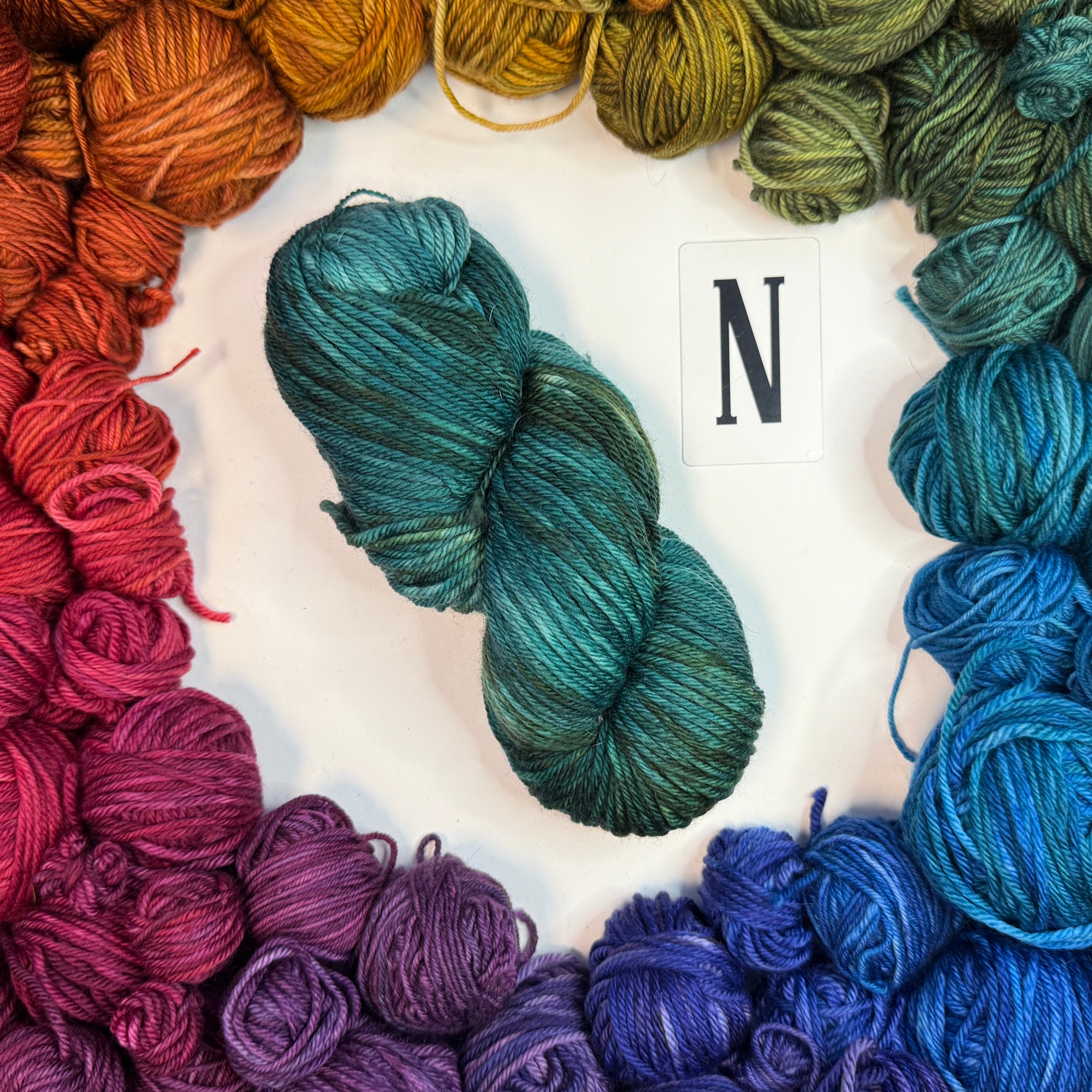 A blue-green skein of yarn labeled "N" framed by a circle of a variety of balls of yarn in rainbow order.