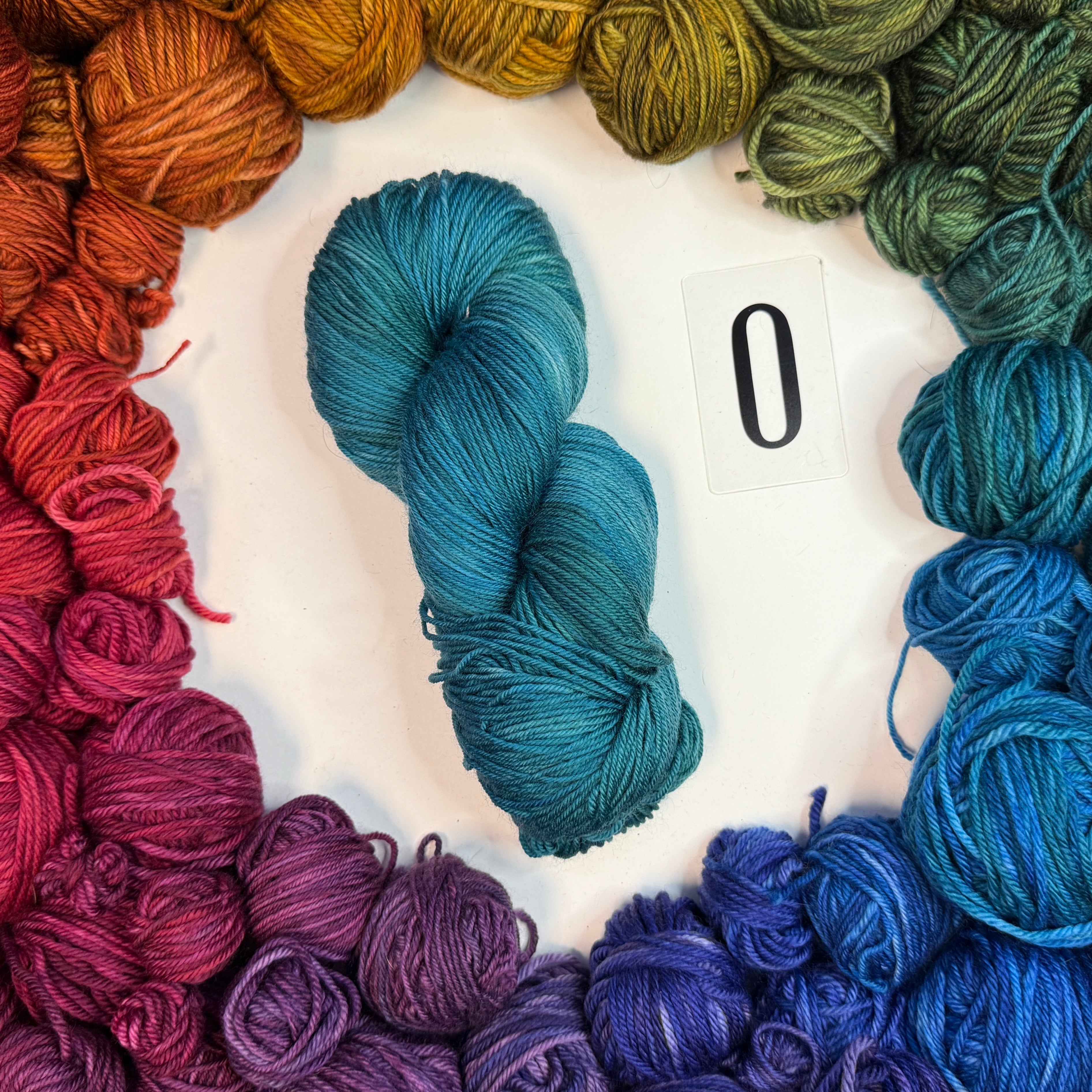 A teal skein of yarn labeled "O" framed by a circle of a variety of balls of yarn in rainbow order.