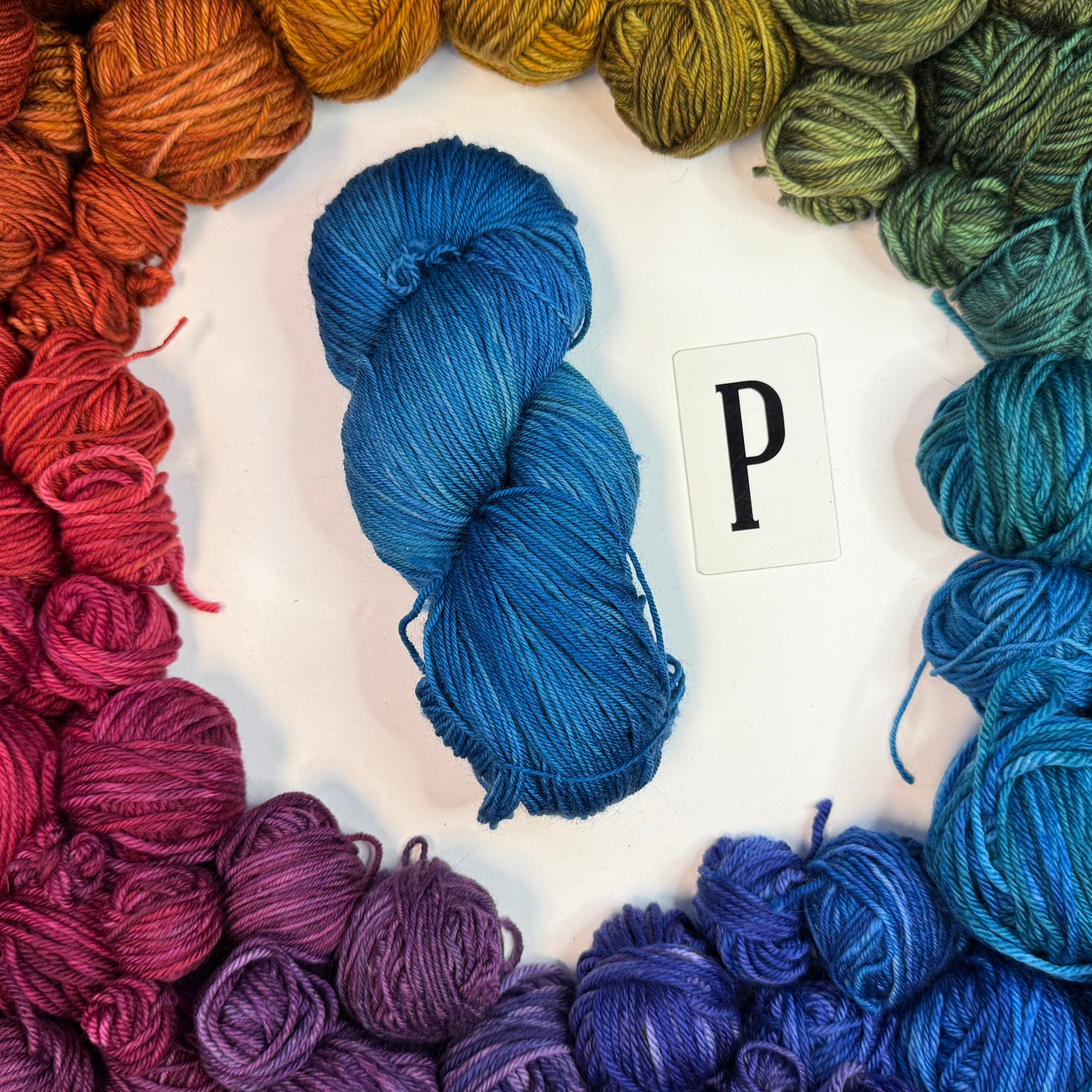 A blue skein of yarn labeled "P" framed by a circle of a variety of balls of yarn in rainbow order.