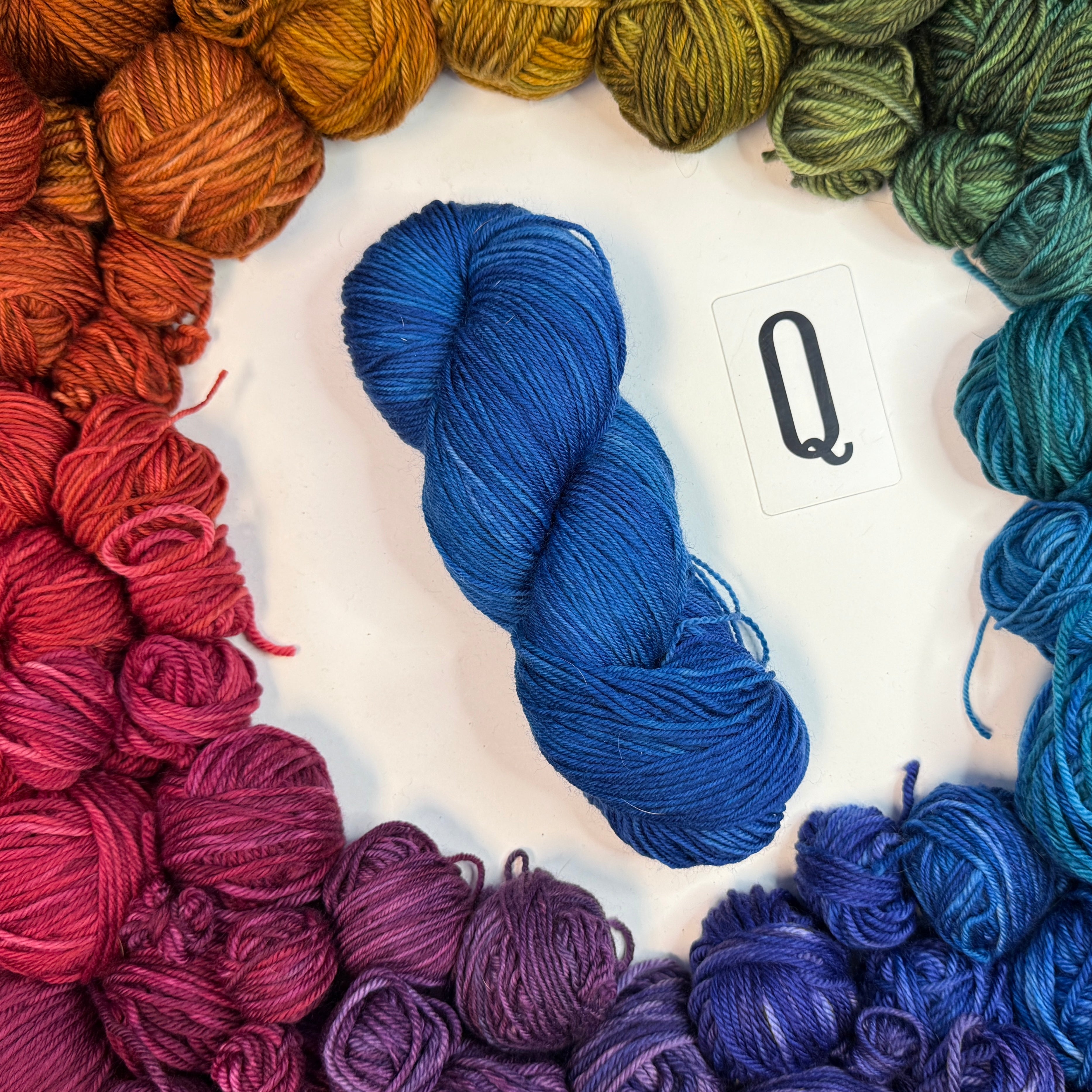 A blue skein of yarn labeled "Q" framed by a circle of a variety of balls of yarn in rainbow order.