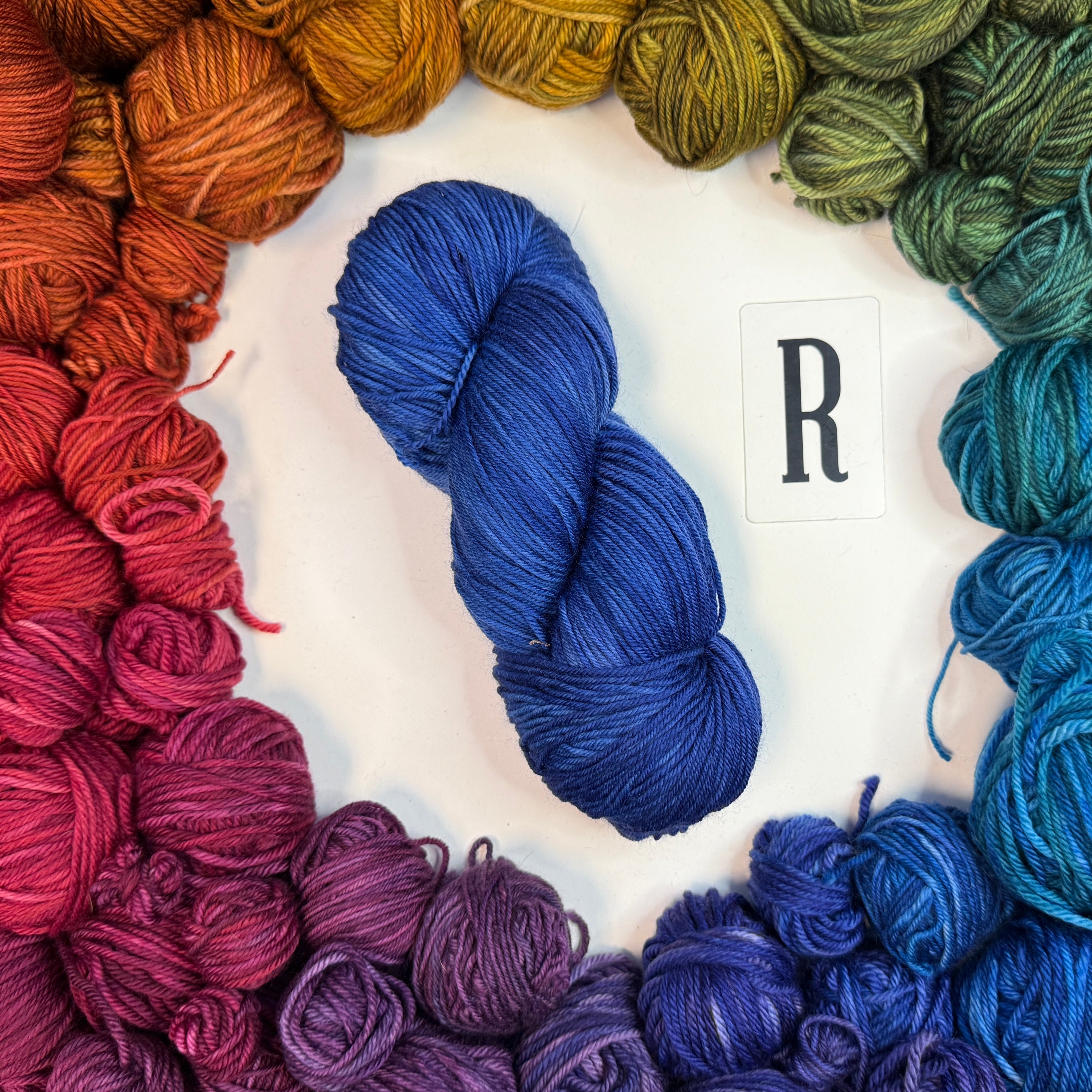 A blue skein of yarn labeled "R" framed by a circle of a variety of balls of yarn in rainbow order.