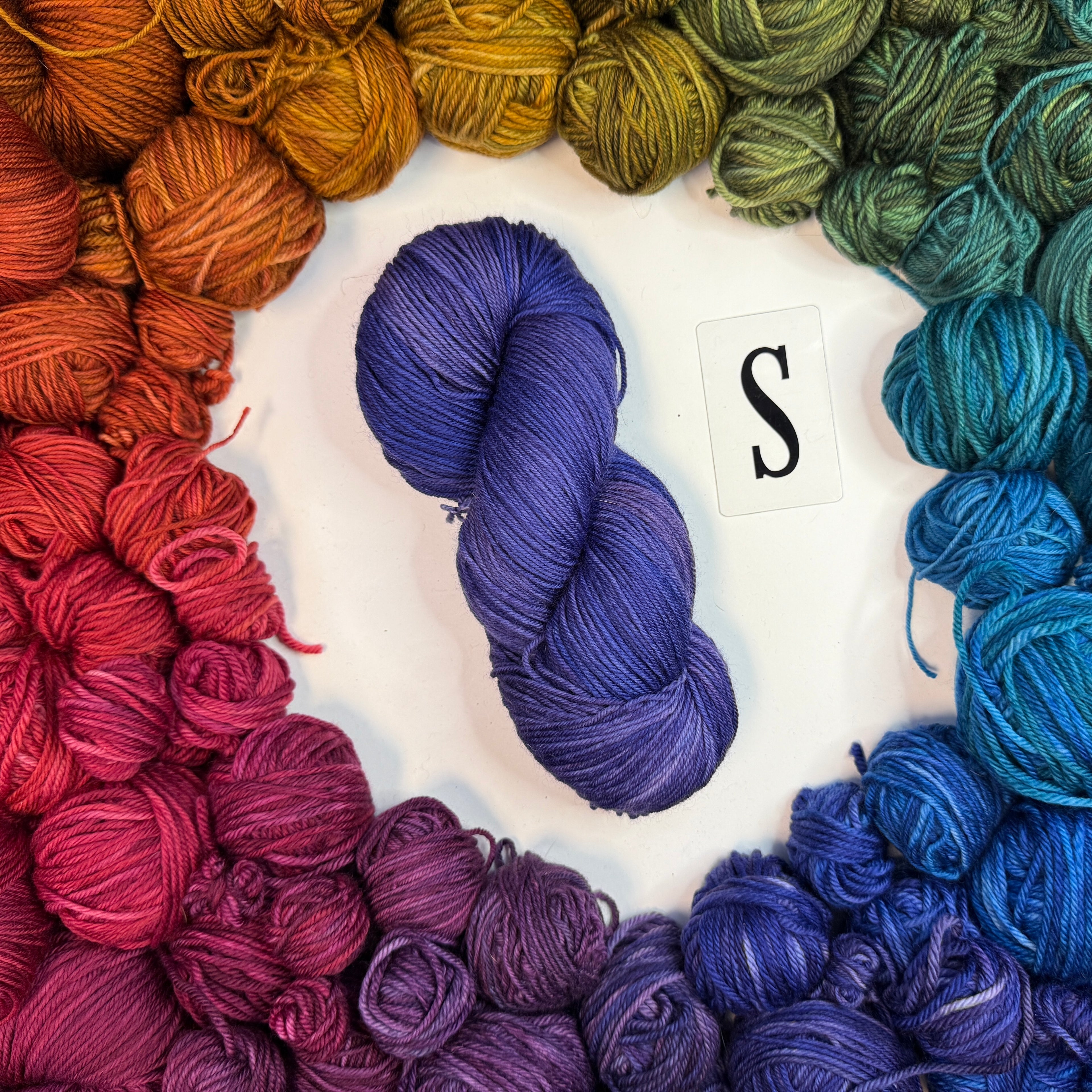 A blue/purple skein of yarn labeled "S" framed by a circle of a variety of balls of yarn in rainbow order.