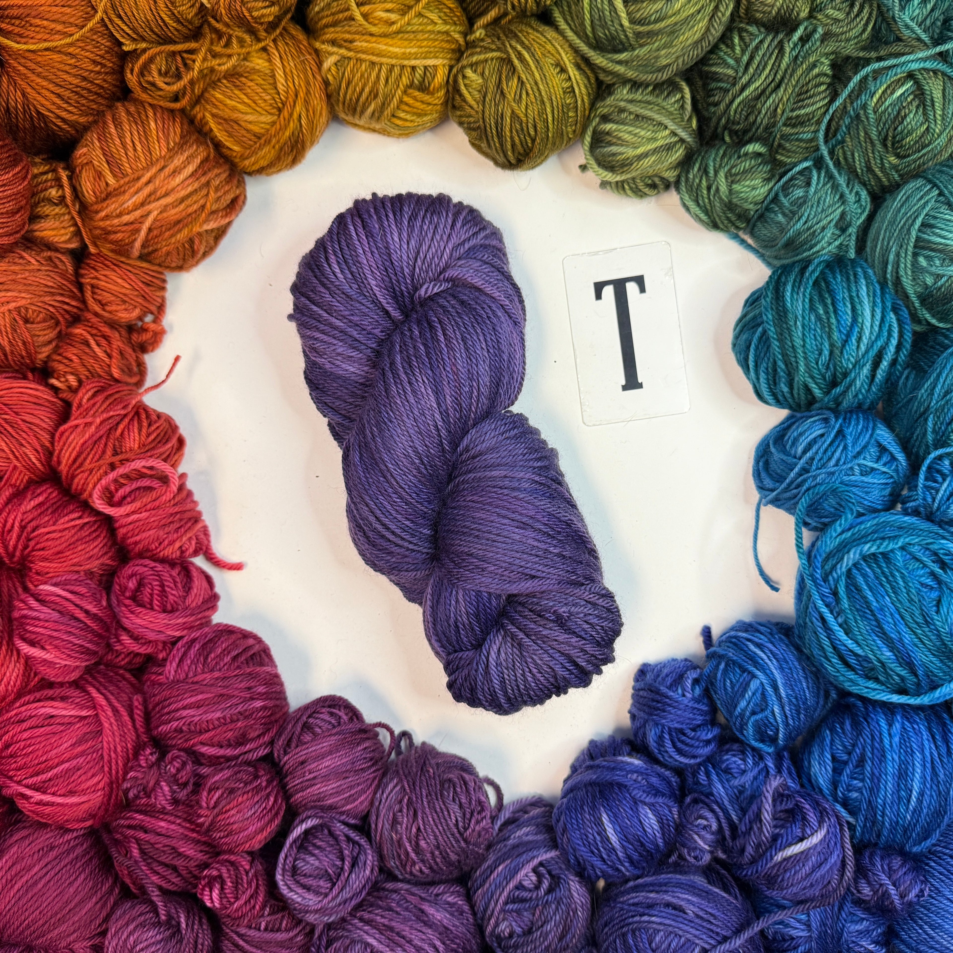 A blue/purple skein of yarn labeled "T" framed by a circle of a variety of balls of yarn in rainbow order.