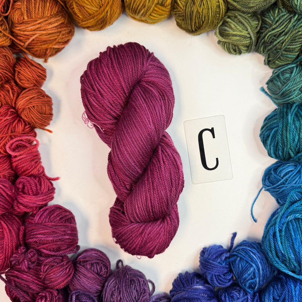 A dark pink skein of yarn labeled "C" framed by a circle of a variety of balls of yarn in rainbow order.