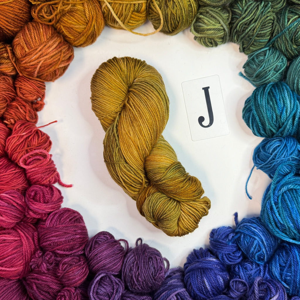 A yellowish skein of yarn labeled "J" framed by a circle of a variety of balls of yarn in rainbow order.